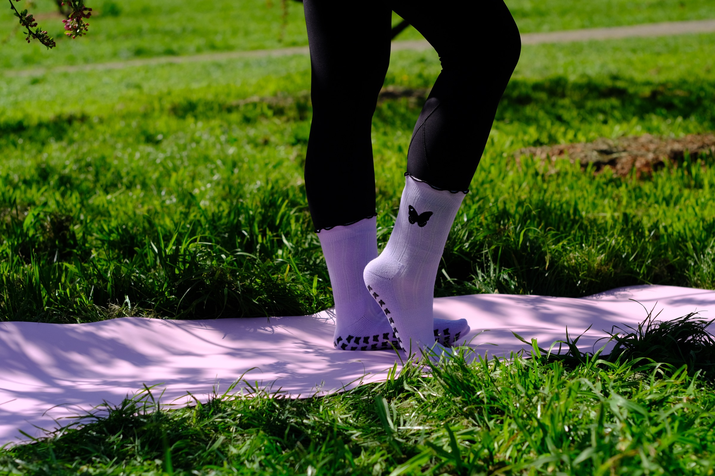 Rebel Butterfly grip socks in lilac, one foot on tiptoe showing the butterfly grip, on a pink mat in the park